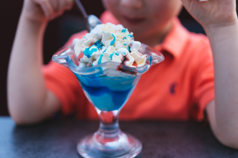 Kind in einem orangefarbenen Poloshirt, das einen Eisbecher mit blauem Sirup und Schlagsahne in einem klassischen Glas genießt.